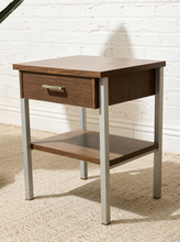 Load image into Gallery viewer, A mid-century modern style nightstand with a dark brown wood finish and silver metal legs, set against a white brick wall and beige carpet.
