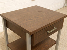 Load image into Gallery viewer, A square wooden nightstand with a medium brown wood-grain finish, featuring a single drawer with a silver metal pull handle and a lower shelf, supported by light gray legs.
