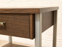Load image into Gallery viewer, Close-up of a mid-century modern wooden desk with a dark brown wood grain finish and a brushed silver metal leg.
