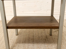 Load image into Gallery viewer, A close-up of a mid-century modern side table featuring a dark brown wood shelf and light gray painted metal legs, set on a woven beige rug against a white brick wall.
