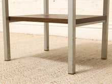 Load image into Gallery viewer, Close-up of a table leg frame with a dark brown wooden shelf and distressed light gray painted legs, set on a beige woven rug against a white brick wall.
