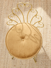 Load image into Gallery viewer, A round tufted velvet stool in a warm beige or champagne color, paired with a decorative gold metal frame backrest, set against a textured beige woven rug.
