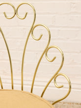 Load image into Gallery viewer, Close-up of a gold metal chair frame with decorative scrollwork and a beige upholstered seat, set against a light brick wall background.
