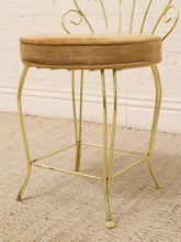 Load image into Gallery viewer, A gold metal stool with a round tan upholstered cushioned seat and a decorative backrest, shown on a woven rug against a white brick wall.
