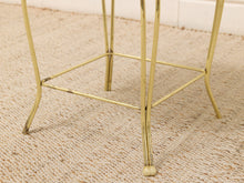 Load image into Gallery viewer, Close-up of a brass metal side table or stool leg frame with a square support brace, standing on a woven beige rug.
