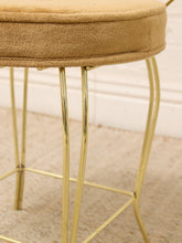 Load image into Gallery viewer, Close-up of a round upholstered stool with a tan textured fabric seat and slim polished gold metal legs, shown in an indoor setting on a light carpet.
