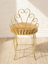 Load image into Gallery viewer, A gold metal barstool with a cushioned tan seat, set against a white brick wall on a woven rug.

