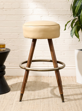 Load image into Gallery viewer, A mid-century modern bar stool with a cream upholstered cushion, dark brown wooden legs, and a brass footrest ring, set against a white brick wall.
