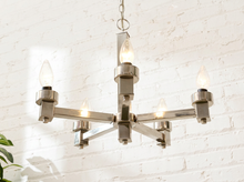 Load image into Gallery viewer, A modern six-light chandelier with a brushed nickel finish and clear glass candle-style bulbs, hanging against a white brick wall.

