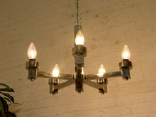 Load image into Gallery viewer, A five-light industrial chandelier with a polished metal frame and glowing light bulbs, hanging against a white brick wall.
