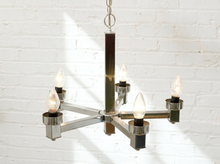 Load image into Gallery viewer, A modern six-light chandelier with a polished silver metal frame and dark rectangular accents, hanging against a white brick wall with warm light from the illuminated bulbs.
