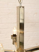 Load image into Gallery viewer, A close-up of a polished silver metal chandelier fixture with a square-shaped vertical stem and a lit bulb, set against a white brick wall background.
