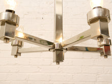 Load image into Gallery viewer, A close-up of a modern multi-light chandelier with a polished chrome finish and clear glass bulbs, shown against a white brick wall background.
