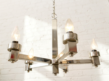Load image into Gallery viewer, A modern six-light chandelier with a polished silver metal frame and clear glass candle-style bulbs, hanging against a white brick wall.
