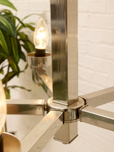 Load image into Gallery viewer, Close-up of a modern metallic chandelier with a brushed silver finish, featuring a central vertical post, horizontal arms, and a lit bulb in a cup holder, set against a white brick wall with a green plant in the background.
