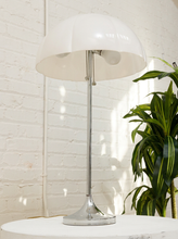 Load image into Gallery viewer, A modern table lamp with a glossy white dome shade and a polished silver metal stem and base, shown on a white table against a white brick wall with a green plant in the background.
