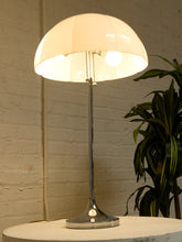 Load image into Gallery viewer, A modern table lamp with a large, dome-shaped off-white shade and a polished silver metal stem and base, shown lit on a round table against a white brick wall with a green plant in the background.
