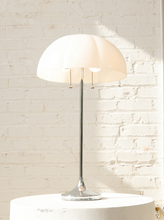 Load image into Gallery viewer, A modern table lamp with a large white dome shade, a slim silver metal stem, and a round silver base, shown on a white pedestal against a white brick wall.
