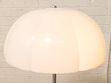Load image into Gallery viewer, A modern table lamp with a large, dome-shaped cream-colored shade made of pleated material, mounted on a slim silver metal stand against a white brick wall background.
