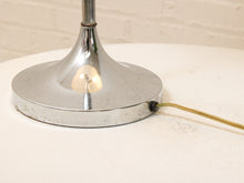 Load image into Gallery viewer, A close-up of a vintage mid-century modern table lamp base with a polished chrome flared collar and a brass-colored cord, shown lit on a white textured surface.
