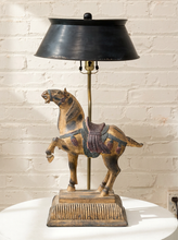 Load image into Gallery viewer, A decorative table lamp featuring a carved horse figurine on a pedestal base with a black metal shade and brass-toned stem, set against a white brick wall.
