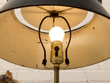Load image into Gallery viewer, Close-up of a vintage-style table lamp with a large black metal shade, a warm yellow interior, a glowing light bulb, and black hanging cords against a white brick wall background.
