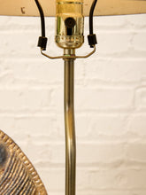 Load image into Gallery viewer, Close-up of a vintage-style table lamp with a curved brass metal stem, a clear glass socket, black hanging cords, and part of a beige lampshade visible against a white brick wall background.
