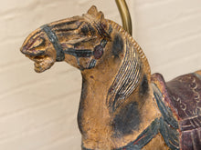 Load image into Gallery viewer, Close-up of a vintage wooden carousel horse head with a weathered gold-brown finish, dark painted details, and a brass-colored hanging loop at the top, set against a light beige brick wall.
