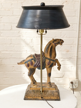 Load image into Gallery viewer, A decorative table lamp featuring a carved horse figurine on a rectangular base, with a black metal shade and brass-toned stem, set against a white brick wall.
