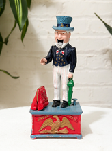 Load image into Gallery viewer, Vintage Uncle Sam figural piggy bank wearing a top hat, dark jacket, bow tie, striped trousers, and standing on a red and blue base
