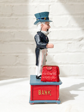 Load image into Gallery viewer, A novelty bobblehead figurine of a man in a top hat and tailcoat standing on a red bankbox with the word 'BANK' in gold, next to a red purse, photographed against a white brick wall.
