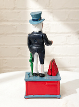 Load image into Gallery viewer, A hand-painted figurine of a man in a black suit, white trousers, and a blue top hat, standing on a red and blue box with a green umbrella and a red bag, photographed against a white brick wall.
