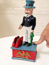 Load image into Gallery viewer, A vintage Uncle Sam toy bank or decorative figurine stands on a red and blue base with an eagle motif. A human hand is touching the side of the figurine, and the background is a white brick wall.
