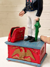 Load image into Gallery viewer, A vintage Uncle Sam toy tin box with a red and blue painted base, gold eagle detail, a red coin purse, and a standing Uncle Sam figure holding a green cane. A human hand is visible holding the figure's leg.
