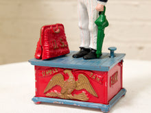 Load image into Gallery viewer, A vintage Uncle Sam toy bank or decorative box with a red body, blue top, gold eagle emblem, and a small red suitcase on top. A partial figure of Uncle Sam stands on the box, wearing white pants and black shoes, against a softly blurred light background.
