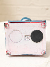 Load image into Gallery viewer, A close-up of the underside of a square object, showing a distressed white painted surface with a circular cutout, a black circular patch, a small screw, and red-painted metal corners on a light fabric background.
