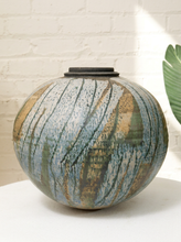 Load image into Gallery viewer, A round ceramic vase with a lid, featuring a marbled pattern of slate blue, green, and earthy brown, displayed on a white surface against a white brick wall.
