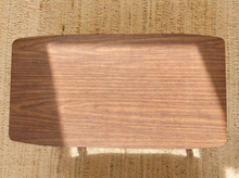 Load image into Gallery viewer, A rectangular wooden table or stool top with rounded corners, shown from above on a woven beige rug. The surface has a warm medium-brown wood grain and is lit by natural light with a soft shadow cast to the left.
