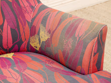 Load image into Gallery viewer, Close-up of upholstered furniture fabric with a bold botanical pattern in coral, pink, red, and purple on a gray base, with a beige carpet visible in the background.
