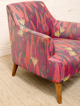 Load image into Gallery viewer, A mid-century modern armchair upholstered in a vibrant pink, red, and orange botanical patterned fabric with light brown wooden legs, set on a woven jute rug against a white brick wall.
