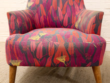 Load image into Gallery viewer, A mid-century modern armchair upholstered in a bold botanical patterned fabric with red, orange, pink, and yellow leaves on a dark background, featuring light wood legs.
