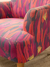 Load image into Gallery viewer, Close-up of a upholstered armchair corner showing a bold botanical pattern in pink, red, orange, and dark gray, with a small portion of a natural wood leg visible on a burlap-textured floor.
