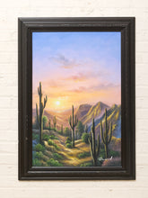 Load image into Gallery viewer, Framed desert sunset landscape painting with tall saguaro cacti, rugged mountains, winding terrain, and colorful golden pink and blue sky

