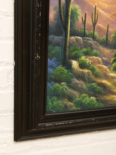 Load image into Gallery viewer, A close-up of a framed desert landscape painting hanging on a white brick wall. The artwork depicts tall saguaro cacti, green shrubs, rocky hills, and warm golden sunlight, enclosed in a dark ornate frame.
