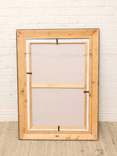 Load image into Gallery viewer, A large, unfinished wooden picture frame with a light natural finish, shown from the back with its canvas and wire hanger frame attached, leaning against a white painted brick wall on a woven rug.
