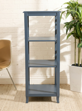 Load image into Gallery viewer, A tall, narrow four-tier shelving unit in a muted slate blue-gray finish, positioned in a styled room setting with a white brick wall, a potted plant, and a partial chair visible.
