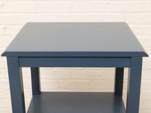 Load image into Gallery viewer, A square side table with a muted blue-gray finish, featuring a top surface and a lower shelf, set against a white painted brick wall.
