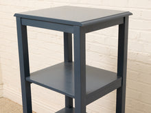 Load image into Gallery viewer, A three-tier wooden shelving unit in a dark slate blue-gray finish, shown against a white brick wall background.
