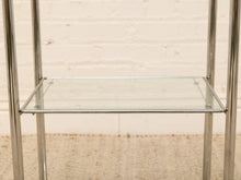 Load image into Gallery viewer, A close-up of a modern shelving unit with a clear glass shelf and polished silver metal legs, set against a light beige brick wall and textured beige rug.
