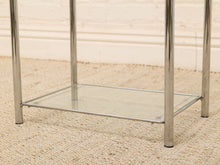 Load image into Gallery viewer, A close-up of a modern side table with a polished chrome metal frame and a clear glass shelf, set on a beige woven rug against a white brick wall.
